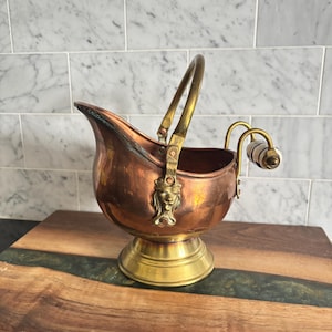 Peut inclure: Un seau à charbon en cuivre avec une base sur piédestal en laiton et une anse en laiton. Le récipient présente une fixation décorative en laiton en forme de tête de lion. Une poignée secondaire à l'arrière comporte une prise en céramique blanche.