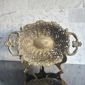 May include: An antique brass-coloured metal serving dish with an elaborate openwork pattern and decorative handles. The oval dish sits on a matching stand, reflecting a vintage aesthetic. The dish is ideal for serving fruit or decorative items.