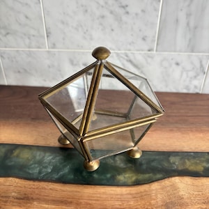 May include: A pentagonal glass trinket box with a brass frame and a pointed lid. The container features five clear glass panels, a central brass finial on top, and stands on three small, rounded brass feet at the base of the structure.