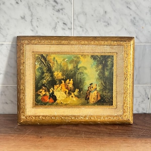 May include: A framed painting showing a pastoral scene with figures in period clothing. The artwork has a gold-coloured ornate frame. The painting's colours include greens, yellows, and browns, with a light blue sky visible in the background.
