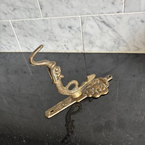 May include: A brass wall hook featuring a sculpted sea creature design with a curved tail and a floral base plate. The metal hardware has a weathered finish and includes two circular mounting holes at the ends of the elongated flat mounting bracket.
