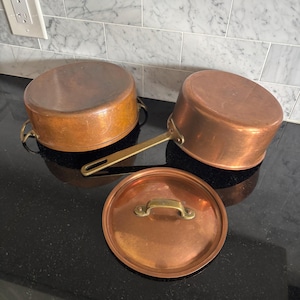 May include: Two round copper pots with metallic handles and a matching circular copper lid. The pots feature a polished finish with visible signs of use, including a long handle on one pot and two side handles on the other, all made of brass.