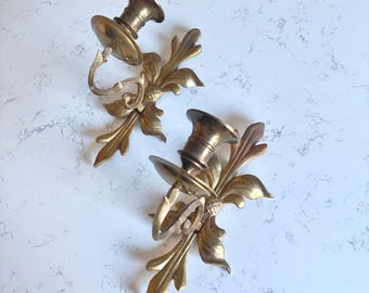 May include: A pair of gold-toned brass wall sconces featuring a central candle holder cup attached to a decorative backplate with ornate leaf and floral motifs. The metal surface has a polished finish with visible metallic reflections.
