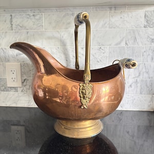 May include: A polished copper coal scuttle with a brass base and a brass handle featuring a lion head mount. The handle includes a white ceramic grip with blue floral patterns. A smaller matching ceramic grip is attached to the rear of the vessel.