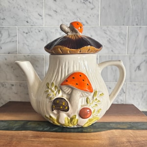 May include: A cream-coloured ceramic teapot featuring a textured, ribbed body and a lid shaped like a brown mushroom cap with a small orange mushroom finial. The front displays raised, hand-painted mushrooms in orange and brown with green foliage.
