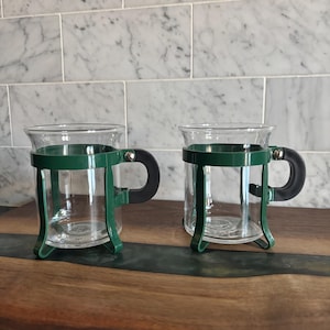 Peut inclure: Deux tasses en verre transparent avec des supports en métal vert foncé et des poignées noires. Les tasses ont un design classique, parfait pour servir des boissons chaudes. Les supports sont fixés avec des rivets argentés.