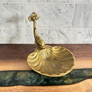 May include: A brass seashell-shaped dish with a decorative dolphin-like stem and a floral top. The dish is a warm gold colour and sits on a wooden surface. The background features a white and grey tiled wall.