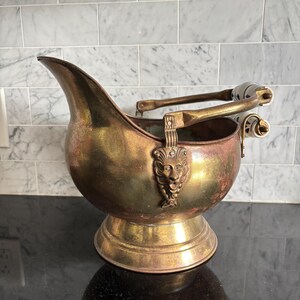 May include: A brass coal scuttle featuring a wide pouring spout, a pedestal base, and a hinged handle with a white ceramic grip decorated with blue floral patterns. The body is adorned with a prominent cast metal lion head relief on each side.