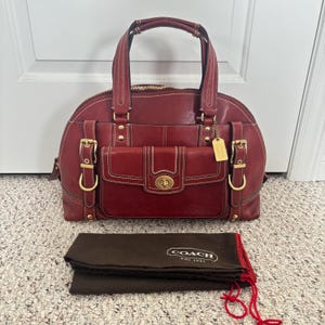 May include: A deep red leather handbag featuring dual top handles, gold-tone hardware, two side buckle straps, and a front pocket with a turn-lock closure. A brown fabric dust bag with the white text COACH EST 1941 is positioned in front of the bag.
