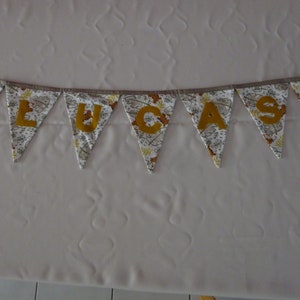 Peut inclure: Une bannière en tissu blanc avec des lettres dorées qui épellent le nom "LUCAS". La bannière est décorée d'un motif de petites feuilles brunes et jaunes.