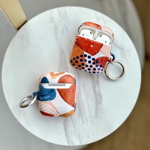 May include: Two AirPod cases with abstract geometric patterns. One case is open, revealing white earbuds inside. Both cases have a silver metal ring attached.