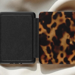 May include: An open black case with a textured finish, next to a cover with a tortoiseshell pattern in shades of brown and tan. The case is designed to protect a device.