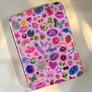 May include: A rectangular pink case covered in a variety of colorful, glittery stickers including rainbows, smiley faces, butterflies, hearts, cherries, a disco ball, a cassette tape, and text reading "girls rule," "too cute," "Angel," and "90% Angel."