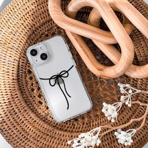 May include: A clear phone case with a black bow design on a woven wicker surface. The phone case is on top of a wooden decorative object.