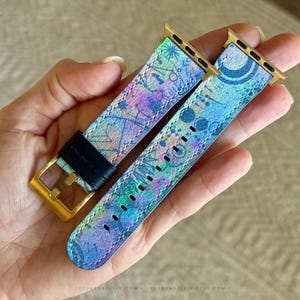 K&ouml;nnte beinhalten: Ein zweiteiliges Uhrenarmband mit einem lebendigen, mehrfarbigen abstrakten Muster in Blau-, Lila- und Gr&uuml;nt&ouml;nen. Das Armband verf&uuml;gt &uuml;ber goldfarbene Metallanschl&uuml;sse und eine goldfarbene Schlie&szlig;e mit einer schwarzen Lederschlaufe.