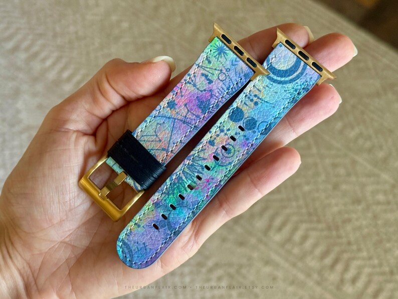 K&ouml;nnte beinhalten: Ein zweiteiliges Uhrenarmband mit einem lebendigen, mehrfarbigen abstrakten Muster in Blau-, Lila- und Gr&uuml;nt&ouml;nen. Das Armband verf&uuml;gt &uuml;ber goldfarbene Metallanschl&uuml;sse und eine goldfarbene Schlie&szlig;e mit einer schwarzen Lederschlaufe.