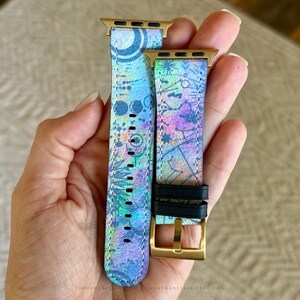 K&ouml;nnte beinhalten: Ein zweiteiliges Uhrenarmband mit einem lebendigen, mehrfarbigen abstrakten Muster in Blau-, Lila- und Gr&uuml;nt&ouml;nen. Das Armband verf&uuml;gt &uuml;ber goldfarbene Metallanschl&uuml;sse und eine goldfarbene Schlie&szlig;e mit zwei schwarzen Lederschlaufen zur Befestigung.