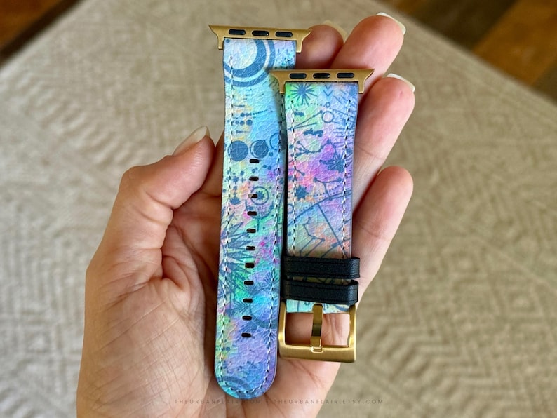 K&ouml;nnte beinhalten: Ein zweiteiliges Uhrenarmband mit einem lebendigen, mehrfarbigen abstrakten Muster in Blau-, Lila- und Gr&uuml;nt&ouml;nen. Das Armband verf&uuml;gt &uuml;ber goldfarbene Metallanschl&uuml;sse und eine goldfarbene Schlie&szlig;e mit zwei schwarzen Lederschlaufen zur Befestigung.