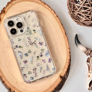 Pode incluir: Uma capa transparente para telefone com um design floral com flores secas e um anel de lente de câmera dourado. A capa está sobre uma superfície de madeira.