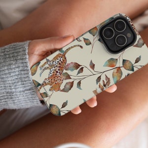 Pu&ograve; includere: Una custodia per iPhone beige con un design ad acquerello di un leopardo e foglie. La custodia ha un ritaglio per l'obiettivo della fotocamera e uno sfondo bianco.
