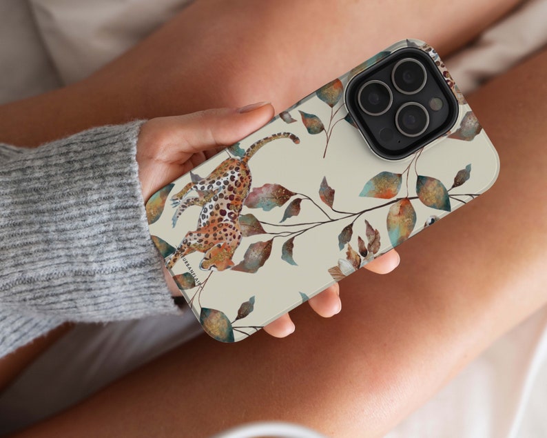 Pu&ograve; includere: Una custodia per iPhone beige con un design ad acquerello di un leopardo e foglie. La custodia ha un ritaglio per l'obiettivo della fotocamera e uno sfondo bianco.