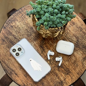 May include: A white iPhone case with a ghost sticker on a wooden table.  A pair of white wireless earbuds and a charging case are also on the table.  A small succulent plant in a gold pot is in the background.