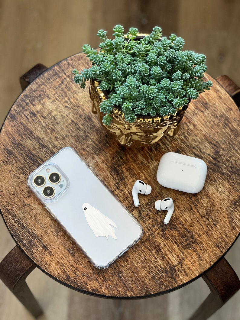 May include: A white iPhone case with a ghost sticker on a wooden table.  A pair of white wireless earbuds and a charging case are also on the table.  A small succulent plant in a gold pot is in the background.