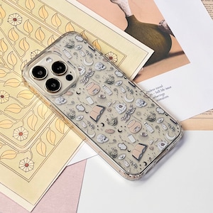 May include: A clear phone case with a white and black pattern of various mystical symbols, including moons, stars, eyes, and hands. The case is designed for an iPhone with a triple camera lens.
