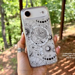 May include: Clear phone case with a celestial design featuring suns, moons, and star charts. The case has a white and black color scheme. The phone case is held in a hand with silver rings. The background is a blurred outdoor scene.