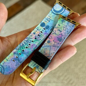 K&ouml;nnte beinhalten: Ein Uhrenarmband mit einem lebendigen, mehrfarbigen abstrakten Muster in Blau-, Gr&uuml;n-, Rosa- und Violettt&ouml;nen. Das Armband verf&uuml;gt &uuml;ber goldfarbene Metallanschl&uuml;sse und eine goldfarbene Schlie&szlig;e, erg&auml;nzt durch eine schwarze Schlaufe.