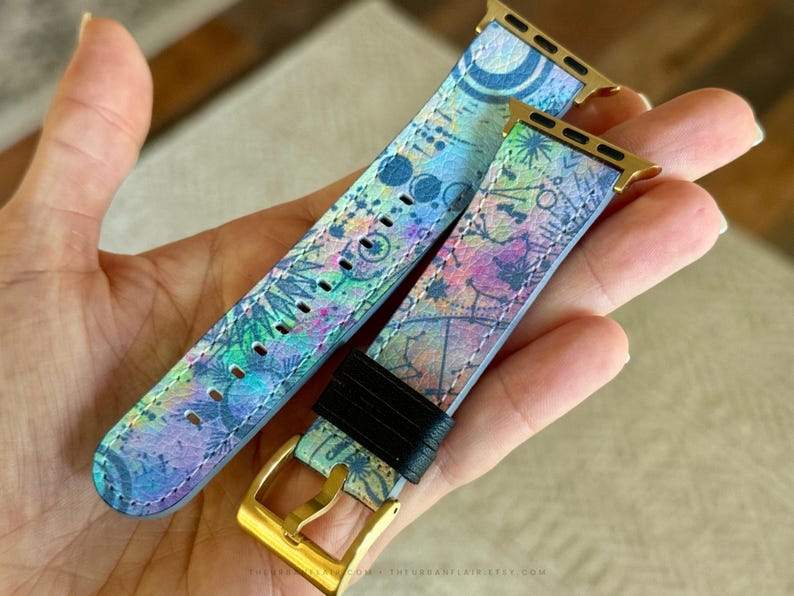 K&ouml;nnte beinhalten: Ein Uhrenarmband mit einem lebendigen, mehrfarbigen abstrakten Muster in Blau-, Gr&uuml;n-, Rosa- und Violettt&ouml;nen. Das Armband verf&uuml;gt &uuml;ber goldfarbene Metallanschl&uuml;sse und eine goldfarbene Schlie&szlig;e, erg&auml;nzt durch eine schwarze Schlaufe.