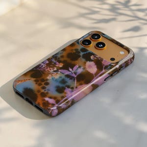 Puede incluir: Una funda para smartphone con un patr&oacute;n tie-dye en tonos marr&oacute;n, azul y morado, con un estampado bot&aacute;nico de hojas y flores. La funda tiene una abertura dorada para el m&oacute;dulo de c&aacute;mara con tres orificios circulares para las lentes.