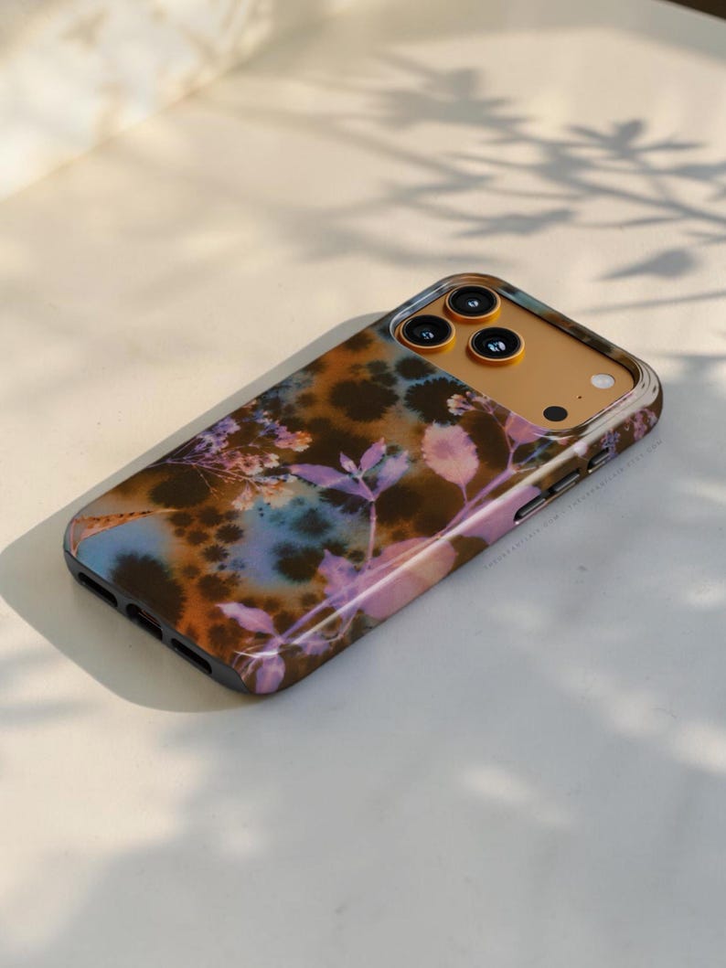 Puede incluir: Una funda para smartphone con un patr&oacute;n tie-dye en tonos marr&oacute;n, azul y morado, con un estampado bot&aacute;nico de hojas y flores. La funda tiene una abertura dorada para el m&oacute;dulo de c&aacute;mara con tres orificios circulares para las lentes.