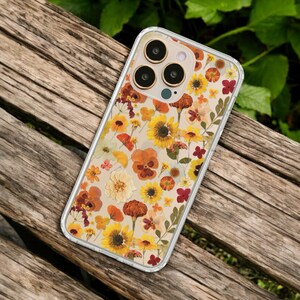 May include: Clear phone case with a floral design featuring pressed yellow sunflowers, orange pansies, and red flowers. The case has a clear border and is set against a wooden background.