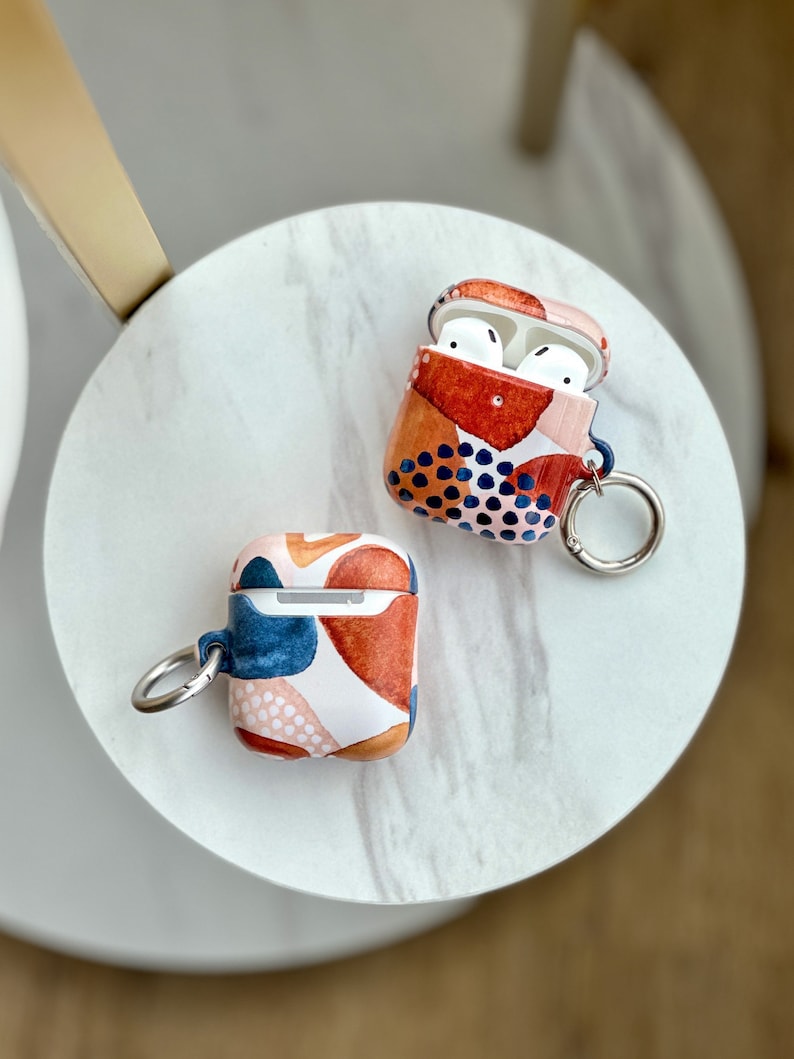May include: Two AirPod cases with abstract geometric patterns. One case is open, revealing white earbuds inside. Both cases have a silver metal ring attached.