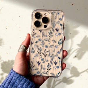 May include: Clear phone case with a floral design featuring blue and white wildflowers. The case has a transparent border and covers the back of the phone. The phone's camera lenses are visible.