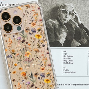 May include: A clear phone case with a floral design. The case features a variety of dried flowers and plants in shades of yellow, orange, purple, and white. The case is designed for a smartphone with a triple camera system.