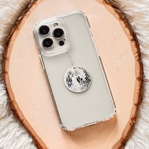 May include: A clear phone case with a silver disco ball charm attached. The phone case is on a wooden surface.