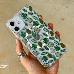 May include: A clear phone case featuring a repeating pattern of various green and beige botanical leaves. The case includes a white circular MagSafe magnetic ring on the back. The text (* MagSafe upgrade pictured) is visible in the bottom left corner.