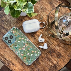 K&ouml;nnte beinhalten: Eine transparente Handyh&uuml;lle mit gr&uuml;nem und wei&szlig;em Blumenmuster, ein wei&szlig;es kabelloses Ladecase mit einem Metallring und zwei wei&szlig;e kabellose Ohrh&ouml;rer auf einer Holzoberfl&auml;che.