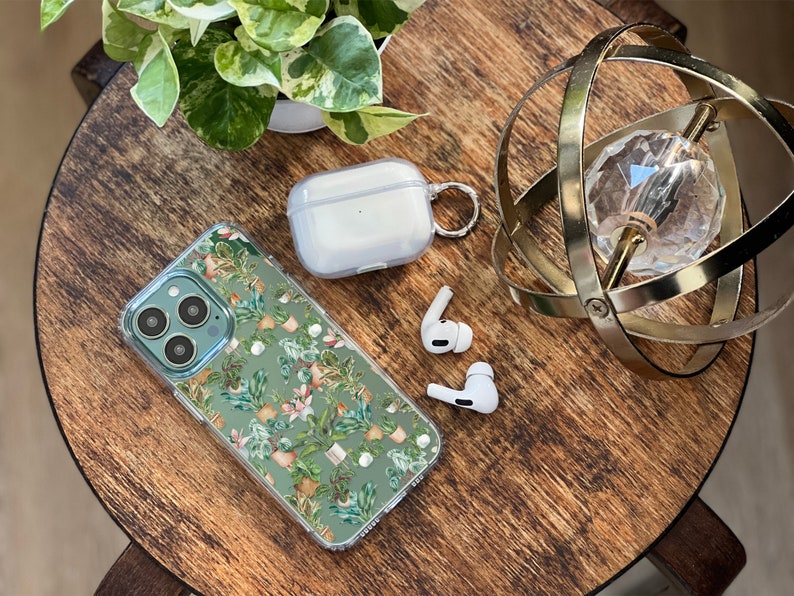 K&ouml;nnte beinhalten: Eine transparente Handyh&uuml;lle mit gr&uuml;nem und wei&szlig;em Blumenmuster, ein wei&szlig;es kabelloses Ladecase mit einem Metallring und zwei wei&szlig;e kabellose Ohrh&ouml;rer auf einer Holzoberfl&auml;che.