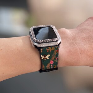 May include: A silver Apple Watch with a clear, bejewelled case and a green floral patterned band with a bee design.