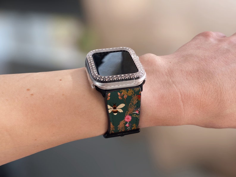 May include: A silver Apple Watch with a clear, bejewelled case and a green floral patterned band with a bee design.