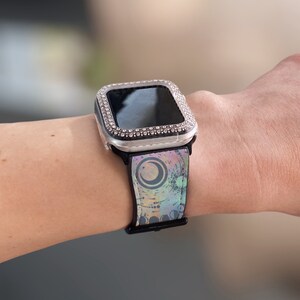 K&ouml;nnte beinhalten: Eine Smartwatch mit einem silberfarbenen Schutzgeh&auml;use, das mit klaren Strasssteinen besetzt ist. Das schwarze Uhrenarmband zeigt einen bunten, abstrakten Grafikdruck mit kreisf&ouml;rmigen Formen und einem Halbmonddesign in Blau- und Violettt&ouml;nen.