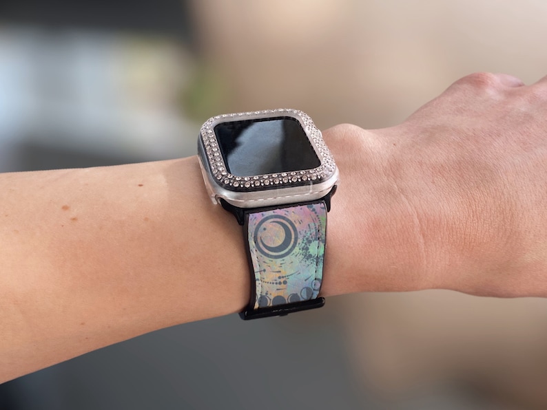 K&ouml;nnte beinhalten: Eine Smartwatch mit einem silberfarbenen Schutzgeh&auml;use, das mit klaren Strasssteinen besetzt ist. Das schwarze Uhrenarmband zeigt einen bunten, abstrakten Grafikdruck mit kreisf&ouml;rmigen Formen und einem Halbmonddesign in Blau- und Violettt&ouml;nen.