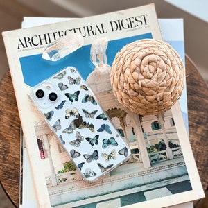 Op de afbeelding: Een transparante telefoonhoes met een vlinderpatroon. De hoes ligt op een stapel tijdschriften, waaronder een exemplaar van Architectural Digest.
