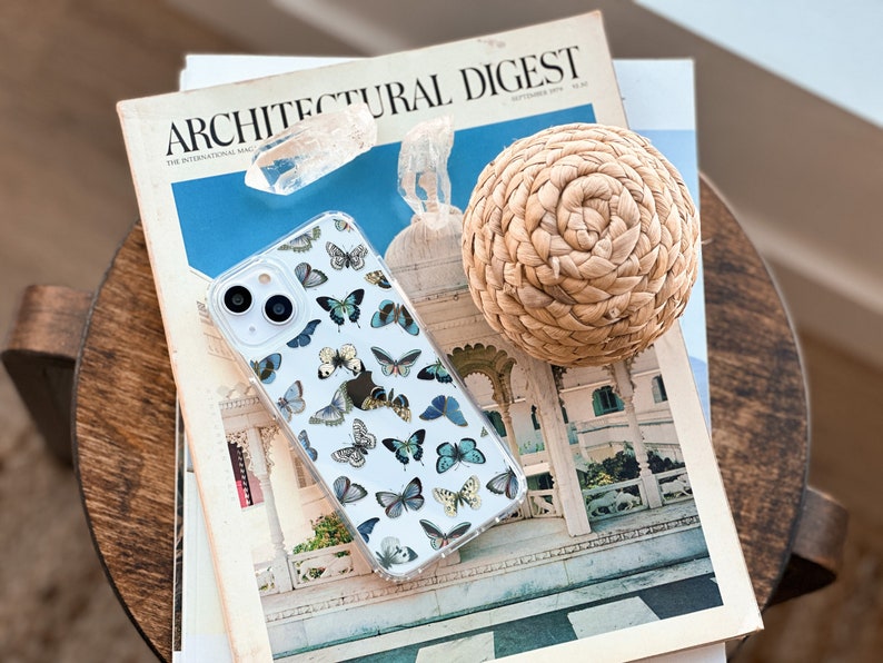 Op de afbeelding: Een transparante telefoonhoes met een vlinderpatroon. De hoes ligt op een stapel tijdschriften, waaronder een exemplaar van Architectural Digest.