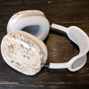 Puede incluir: Auriculares blancos de diadema con un diseño floral en las orejeras. Los auriculares tienen una diadema y almohadillas blancas. El estampado floral presenta elementos botánicos marrones, beige y verdes. Los auriculares están sobre una superficie de madera oscura.