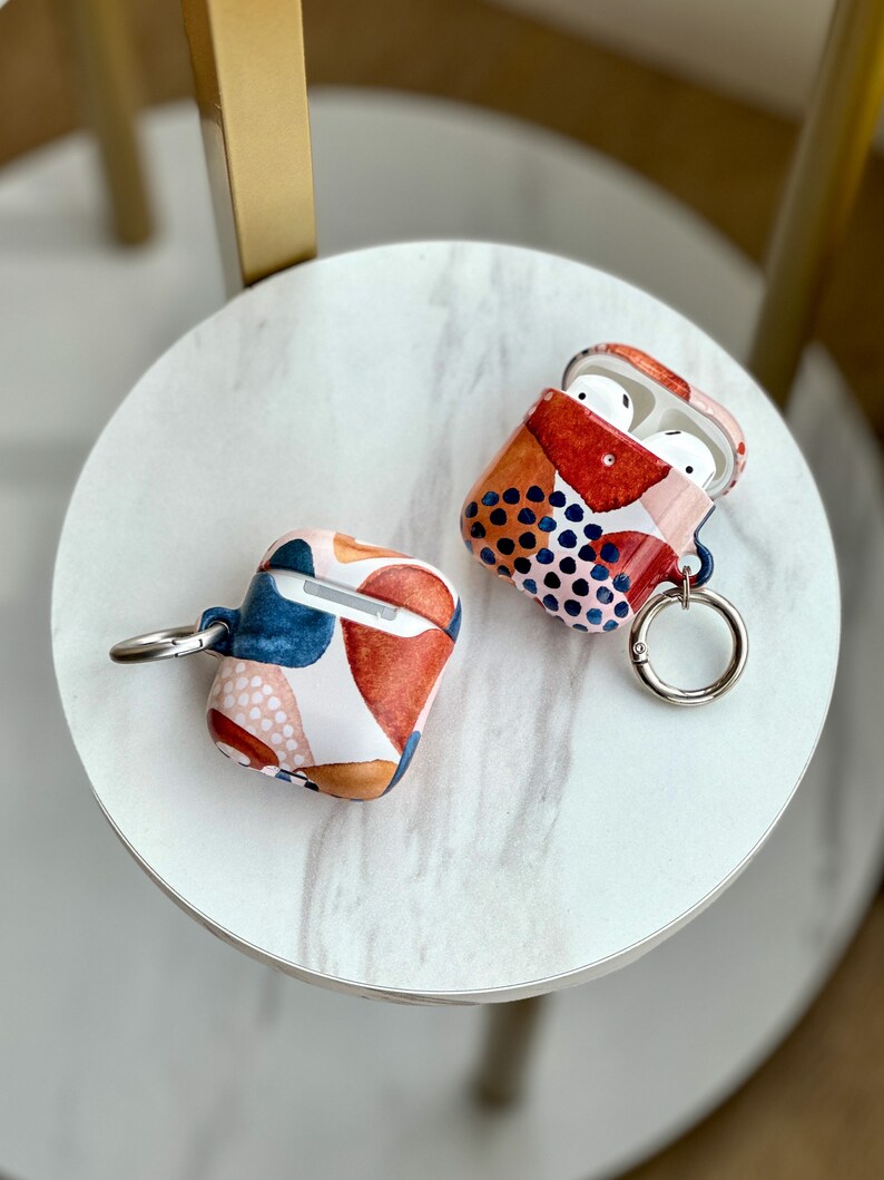 May include: Two Airpod cases with abstract patterns. One case is red, blue, and white with black polka dots. The other case is pink, blue, and orange with white polka dots. Both cases have a silver ring attached.