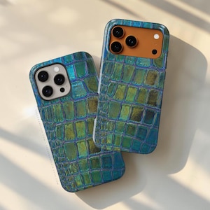 Puede incluir: Dos fundas para smartphone con un patrón texturizado de piel de cocodrilo iridiscente en tonos azul, verde y dorado. Una funda tiene un recorte de cámara claro, mientras que la otra presenta un módulo de cámara beige. Ambas muestran el texto THEURBANFLAIR.COM.